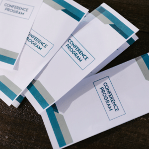 Brochure promozionali impilate su tavolo in legno con grafica bianca e bande turchesi e scritta "Conference Program" visibile