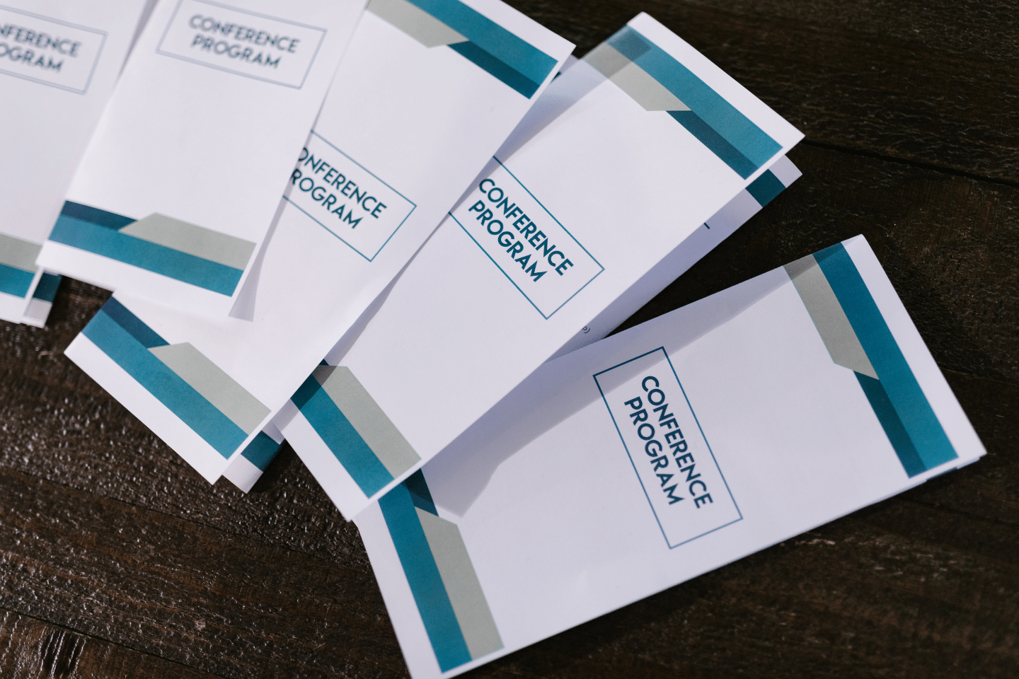 Brochure promozionali impilate su tavolo in legno con grafica bianca e bande turchesi e scritta "Conference Program" visibile