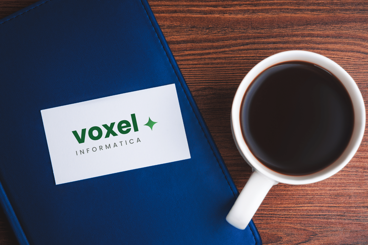 Biglietto da visita Voxel Informatica posato su copertina blu vicino a tazza di caffè su tavolo in legno